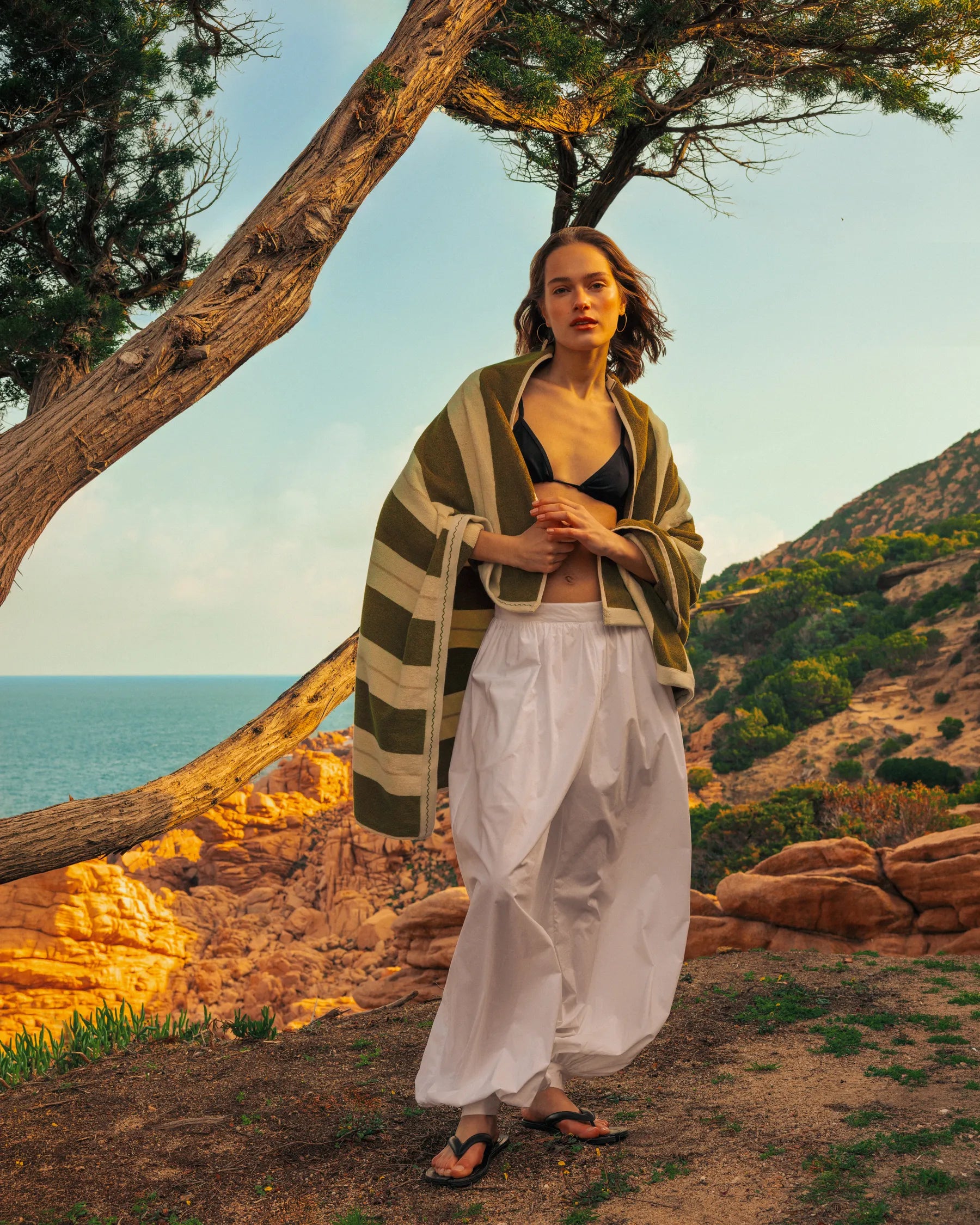 Woman wrapped in Éteanne striped beach towel in Terre Verte standing by Mediterranean coastline
