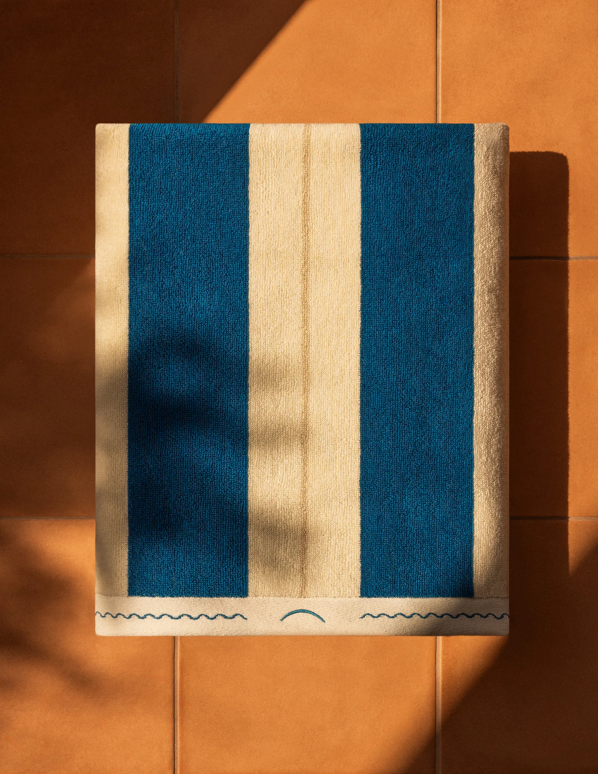 Folded Éteanne Anné striped beach towel in Ora Blu on terracotta floor.