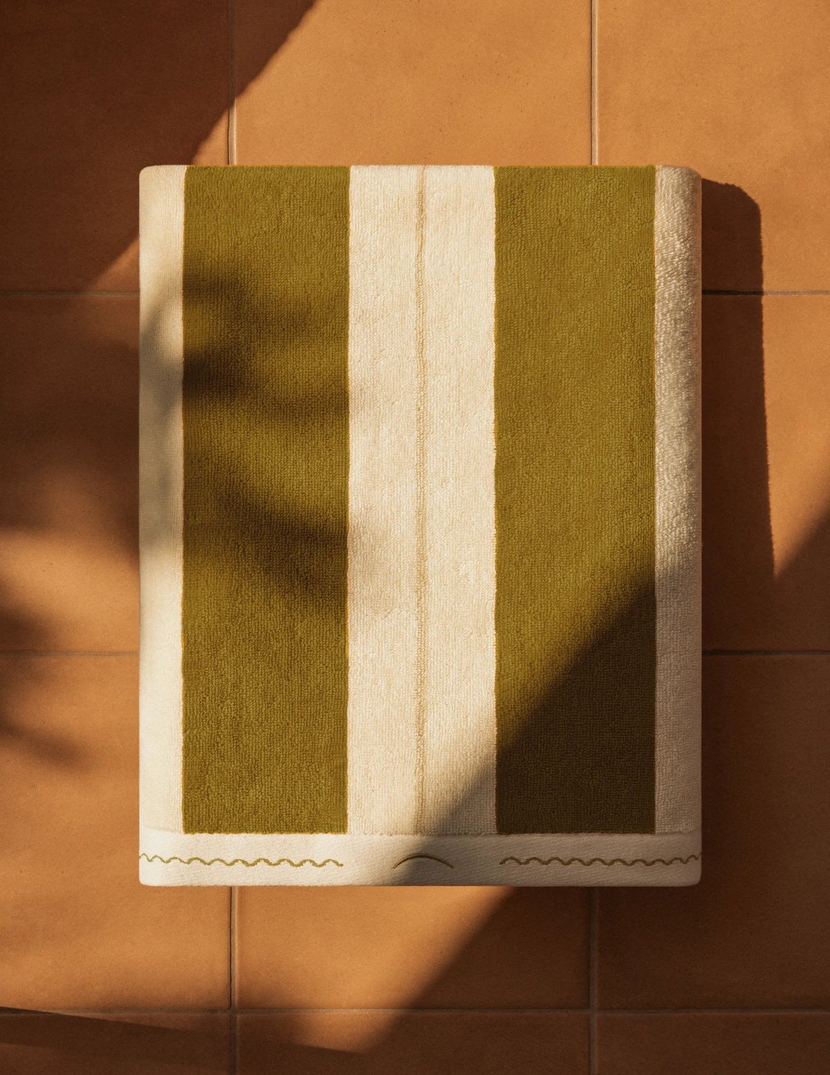 Folded Éteanne Anné striped beach towel in Terre Verte on terracotta floor.