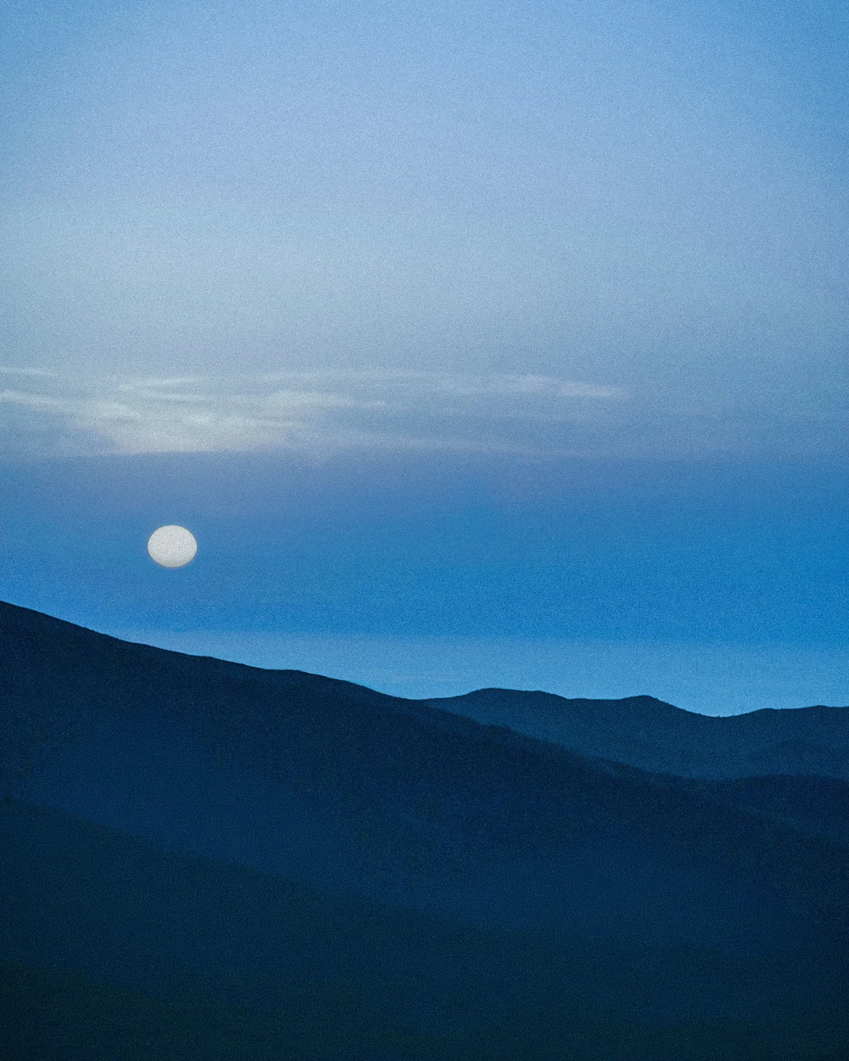 Moon rising above layered mountain landscape at dusk