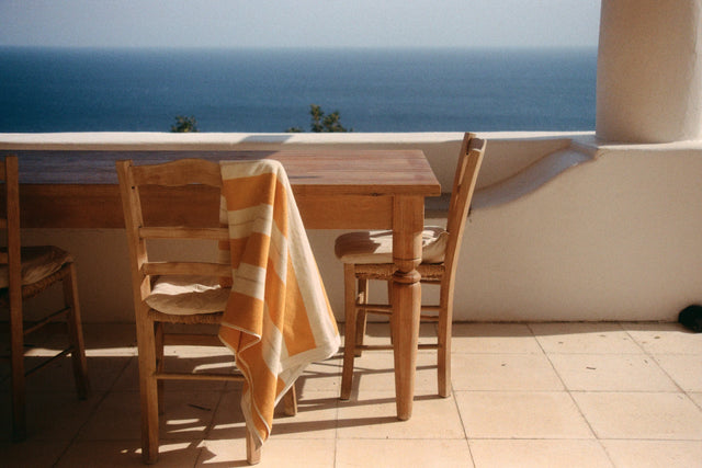 Anne Beach Towel in Tuscan Sun draped over a wooden chair on a Mediterranean terrace overlooking the sea.