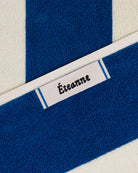 Éteanne woven label embroidered onto striped cotton beach towel in Ora Blu color