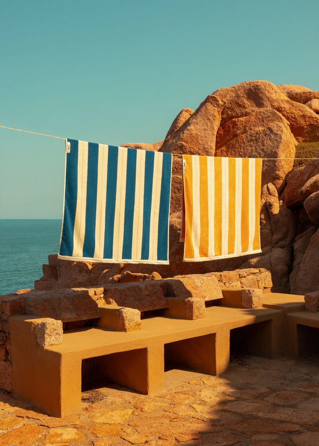 Éteanne striped beach towels in Ora Blu and Tuscan Sun hanging outdoors by rocky coastline