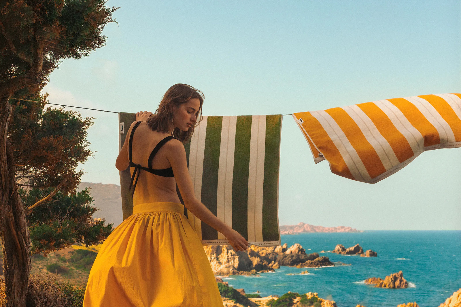 Women hanging Éteanne striped beach towels in Terre Verte and Tuscan Sun hanging outdoors by rocky coastline